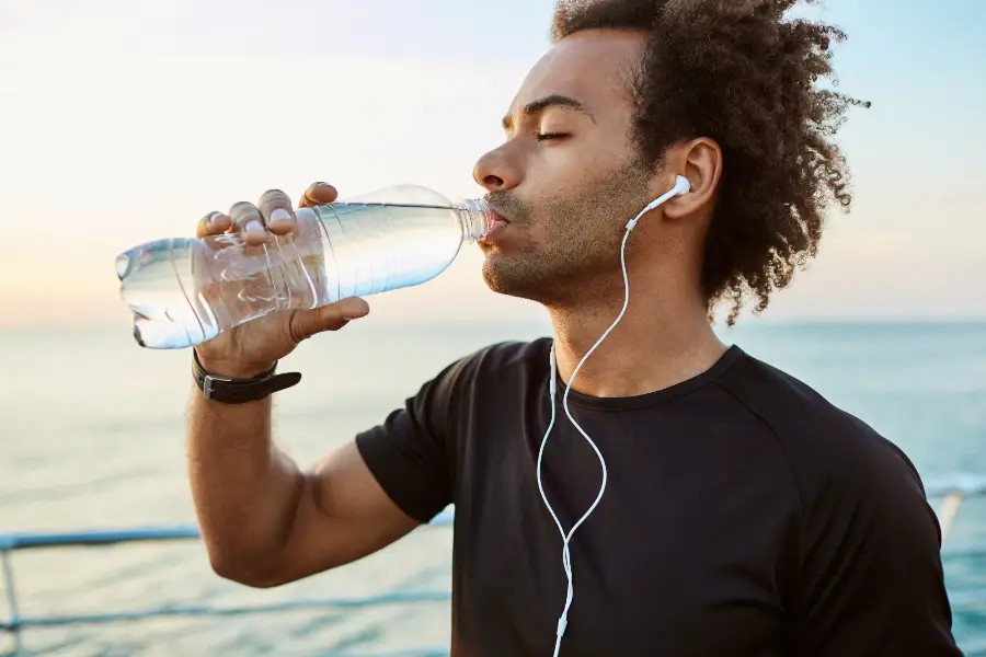 Close up portrait of Afro-American fit athlete drinking water out of plastic bottle with earphones on. Refreshing himself with water and wearing black t-shirt